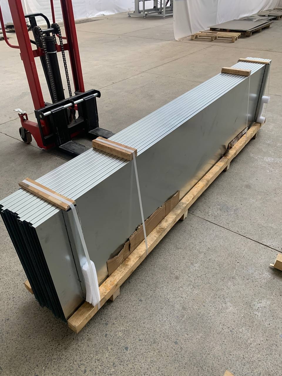 Paczka metalowych paneli na palecie, zabezpieczona drewnianymi klockami i folią, transportowana wózkiem widłowym w hali. Widoczne detale mocowania i struktura paneli.