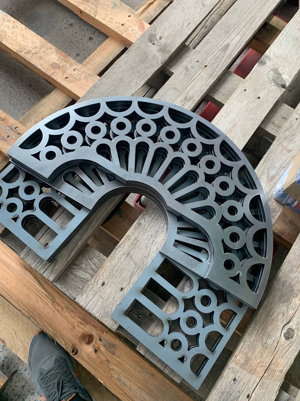 Wycięte laserowo, metalowe elementy dekoracyjne z powtarzalnym wzorem geometrycznym, ułożone na drewnianej palecie. Widoczny fragment buta dla skali.