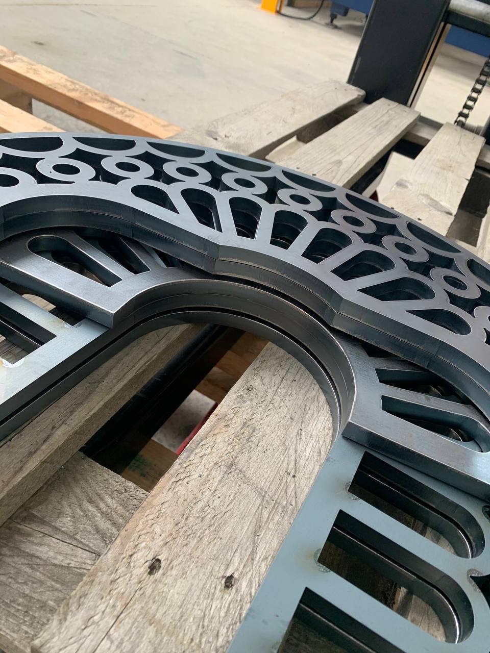 Metalowe elementy z wyciętymi wzorami, ułożone na drewnianej palecie. Precyzyjne cięcie, powtarzalny wzór, surowy materiał. Detal obróbki metalu.