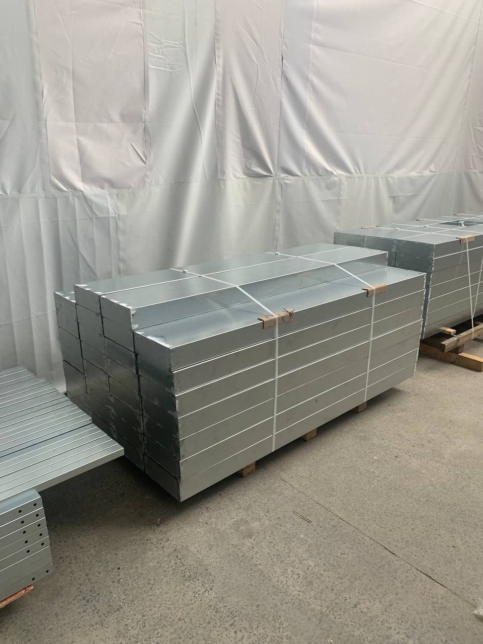 Stosy metalowych elementów, prawdopodobnie do ogrodzeń, zabezpieczone taśmami na drewnianych paletach w hali produkcyjnej. Widoczne otwory montażowe.