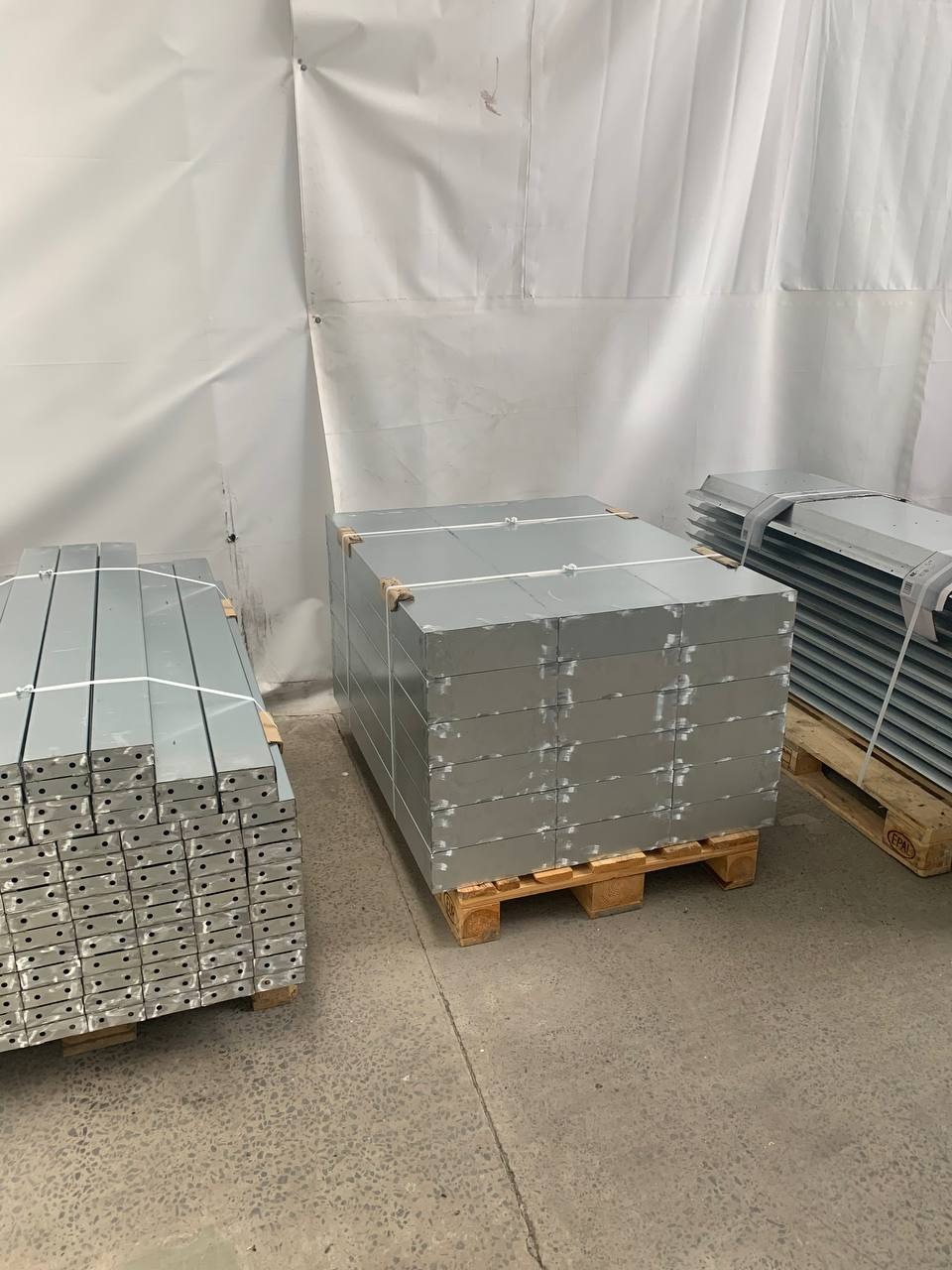 Stosy metalowych elementów na paletach, przygotowane do dalszej obróbki lub transportu. Widoczne otwory montażowe i szare wykończenie.