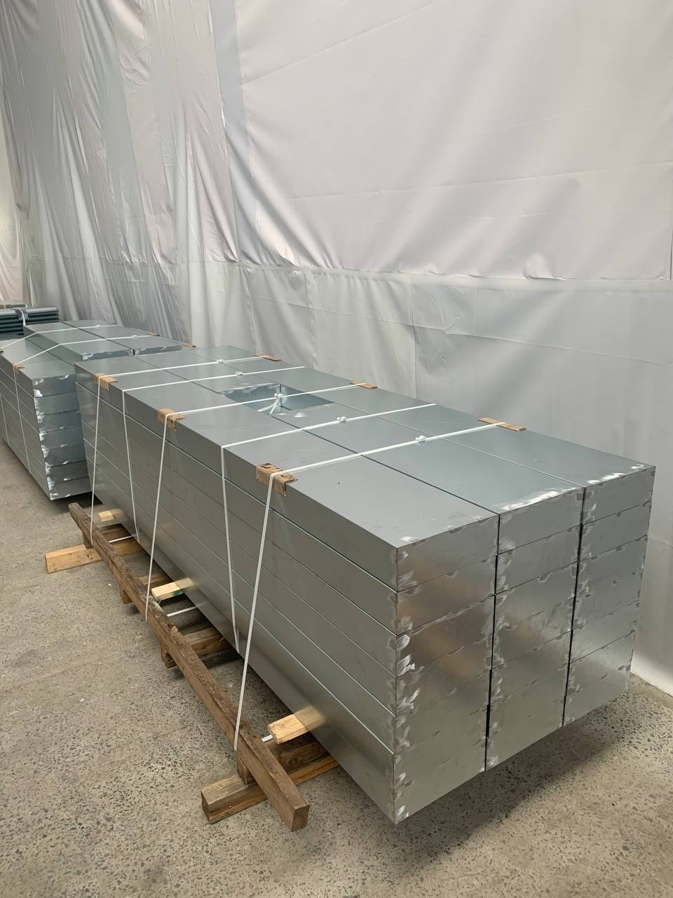 Stosy metalowych elementów, prawdopodobnie blach, ułożone na drewnianych paletach w magazynie. Elementy zabezpieczone białym sznurkiem.