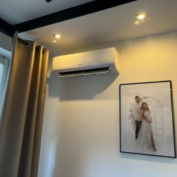 Łukasz Kusztykiewicz KMK klimatyzacja - Biały klimatyzator Hisense na ścianie w jasnym pokoju, obok okna z żaluzjami i oprawionego zdjęcia. Widoczne oświetlenie sufitowe.