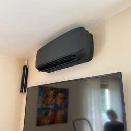 Łukasz Kusztykiewicz KMK klimatyzacja - Czarny klimatyzator ścienny w nowoczesnym wnętrzu, zamontowany nad telewizorem. Widoczny fragment głośnika i obraz na ścianie. Minimalistyczny design.