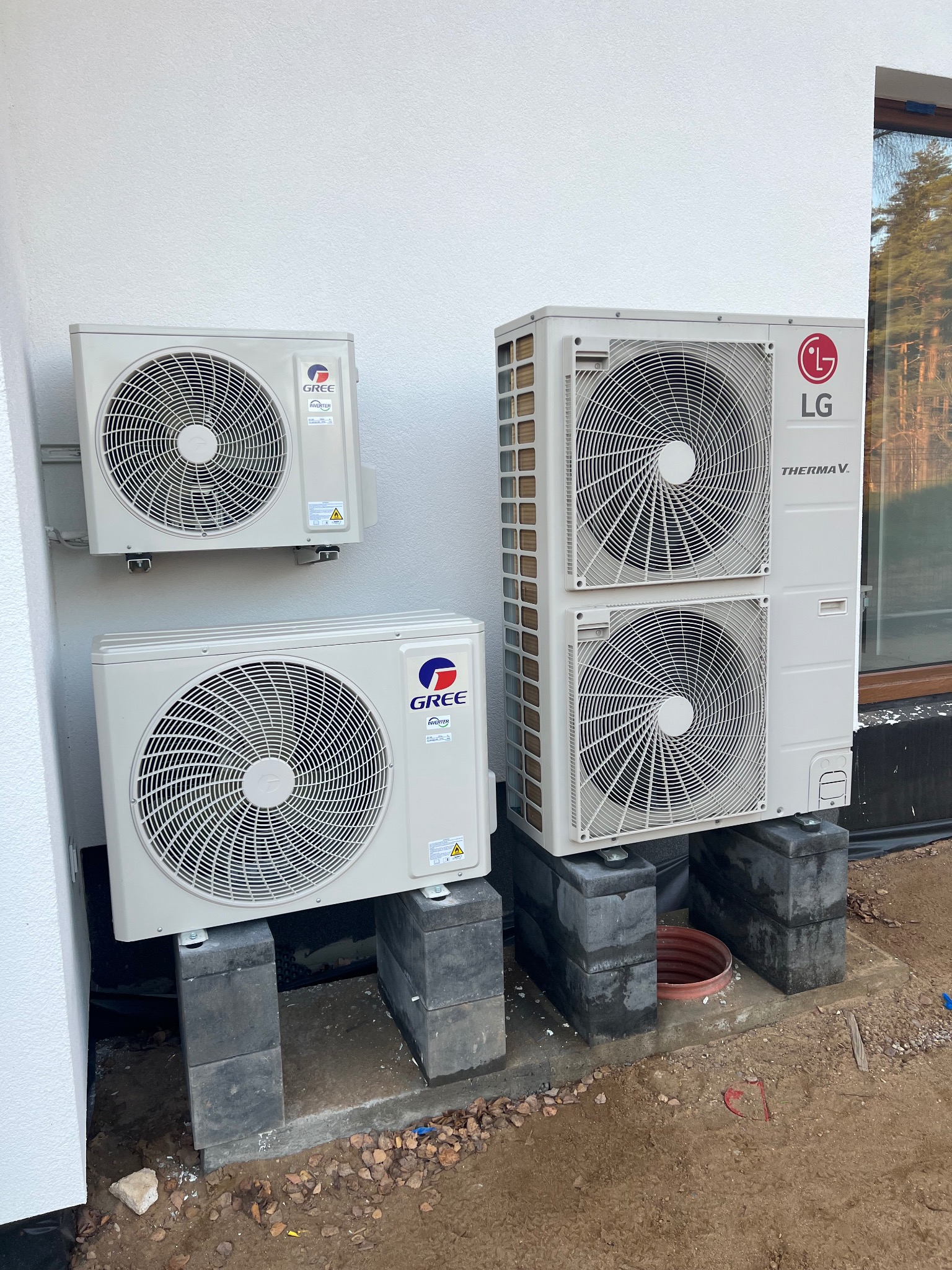 Zewnętrzne jednostki klimatyzacji Gree i LG Thermav V zamontowane na betonowych bloczkach przy białej ścianie budynku. Widoczne elementy instalacji.