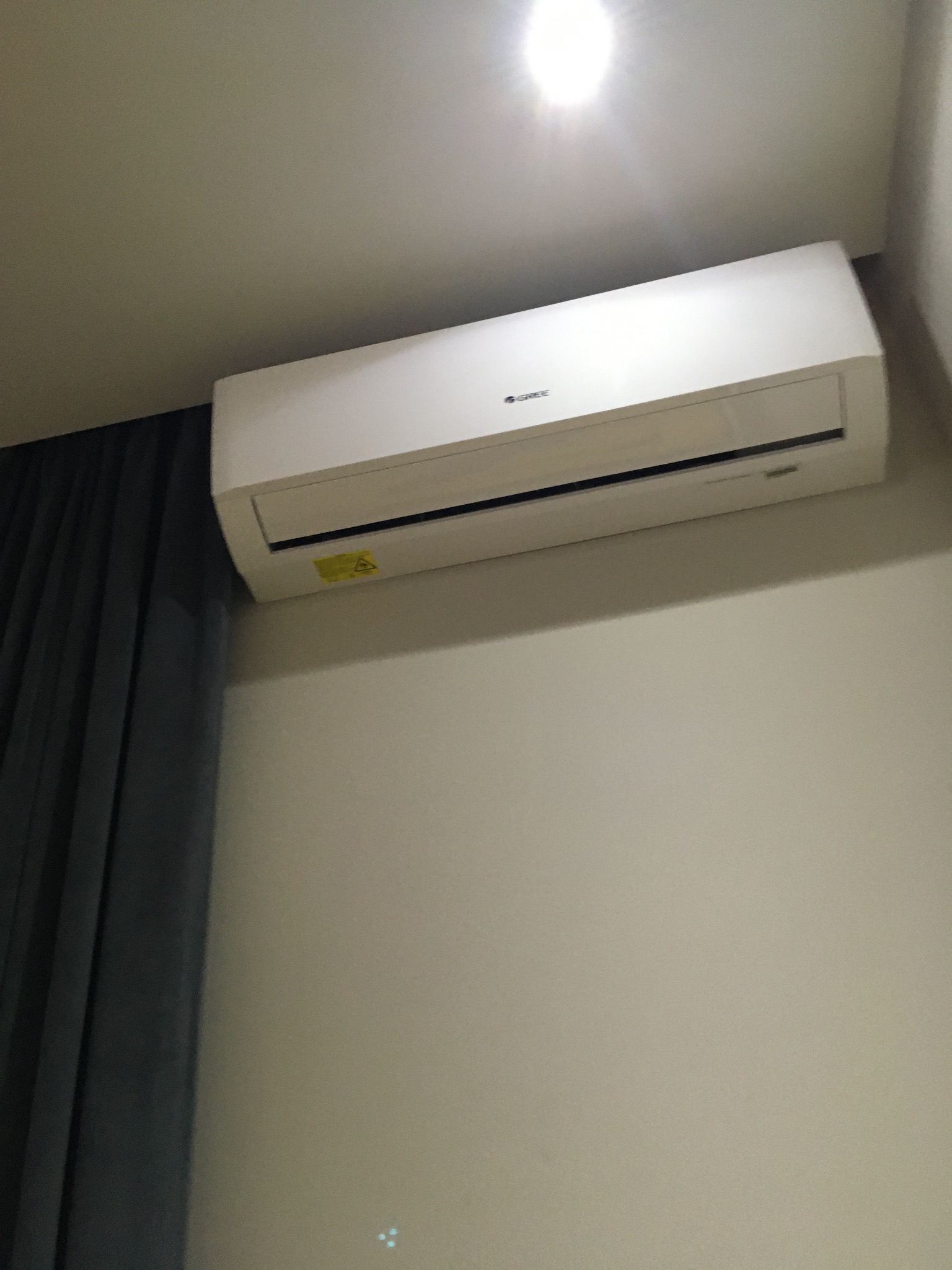 Biały klimatyzator marki Gree zamontowany na ścianie, widoczny fragment zasłony. Oświetlenie sufitowe podkreśla czystość i nowoczesny design urządzenia.