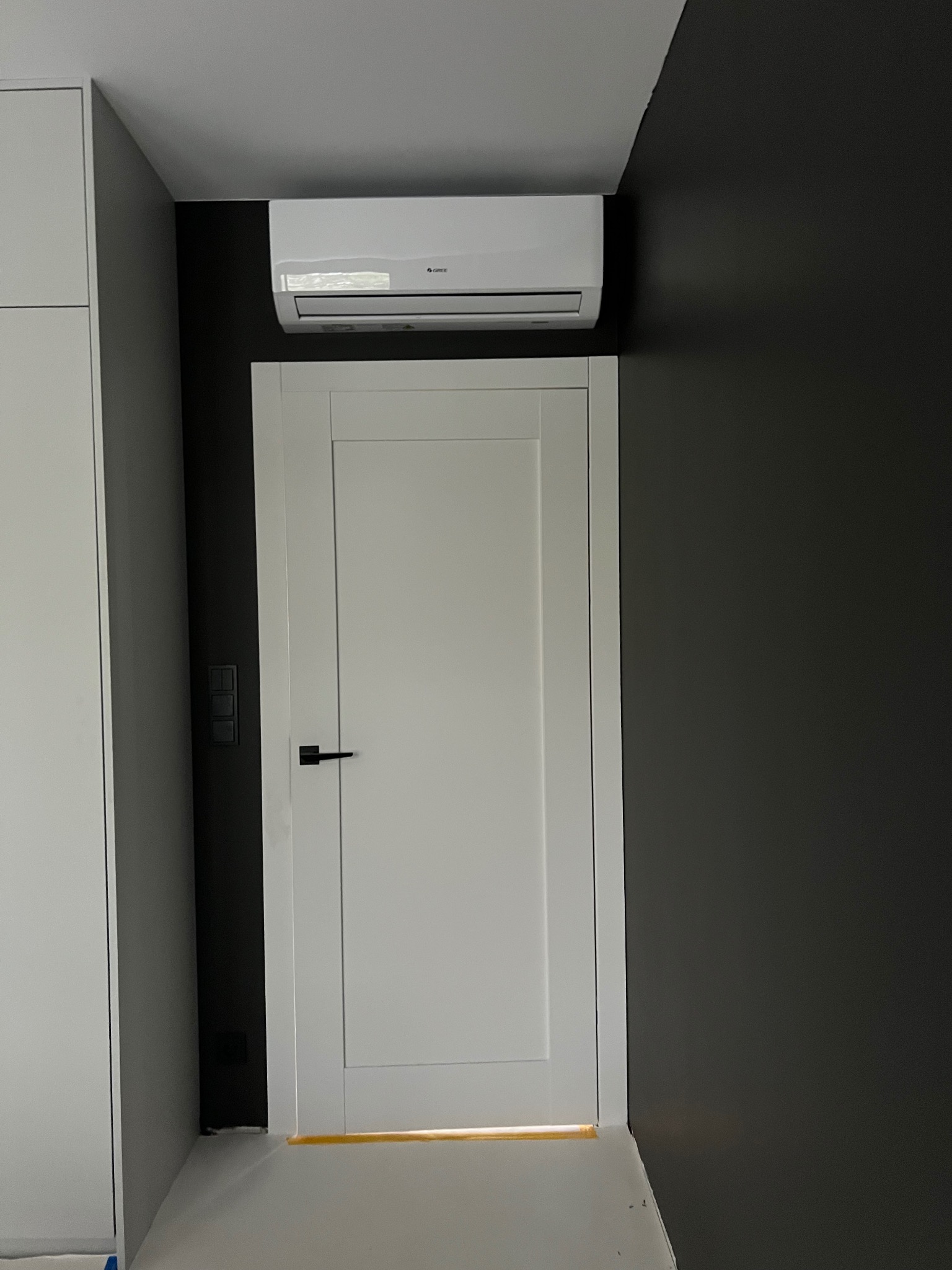 Biały klimatyzator marki Gree zamontowany nad białą, nowoczesną ościeżnicą drzwiową na tle grafitowej ściany w minimalistycznym wnętrzu.