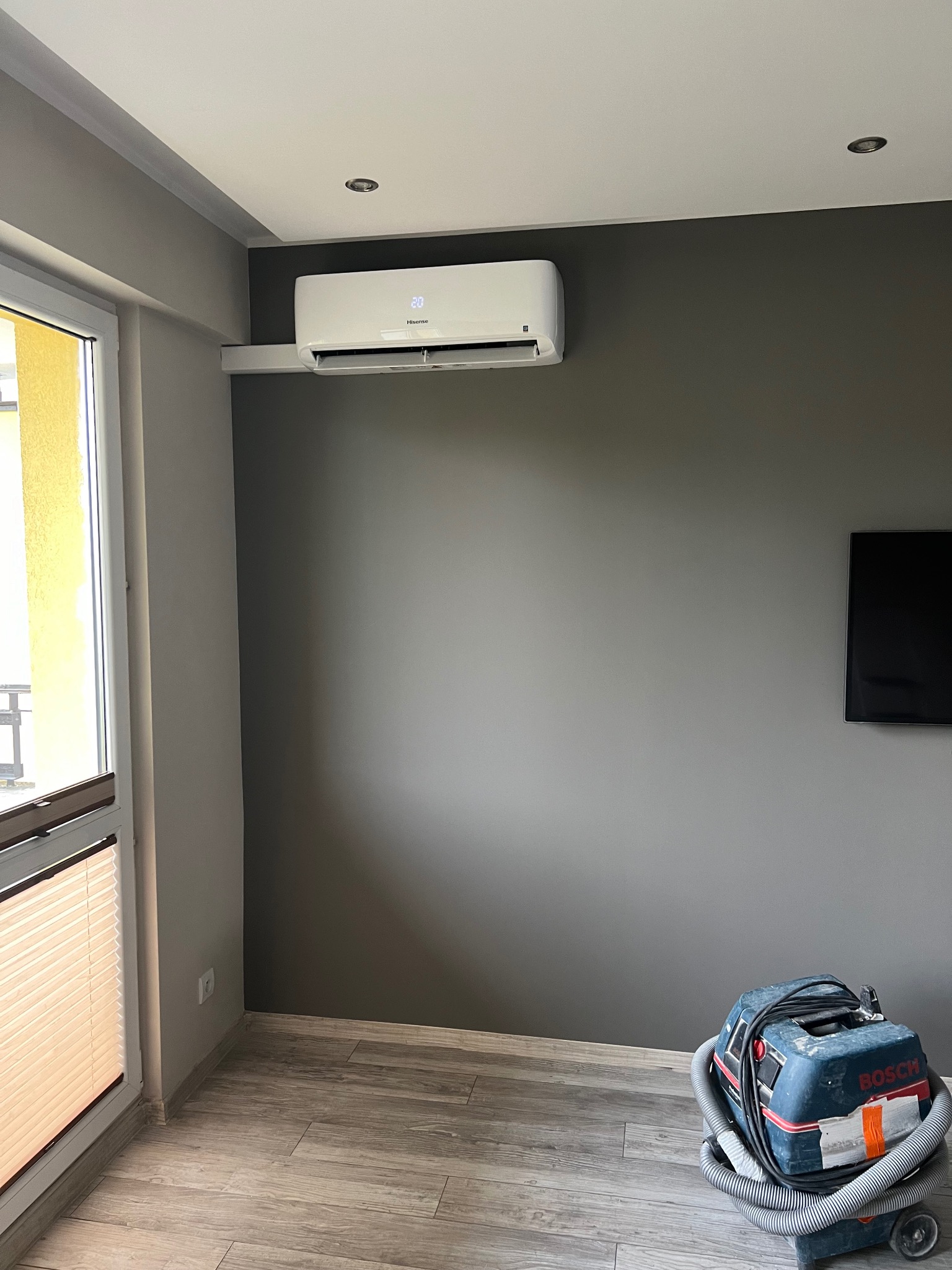 Biały klimatyzator na szarej ścianie w pokoju, widoczny odkurzacz przemysłowy na podłodze. Nowoczesne wnętrze po montażu klimatyzacji.