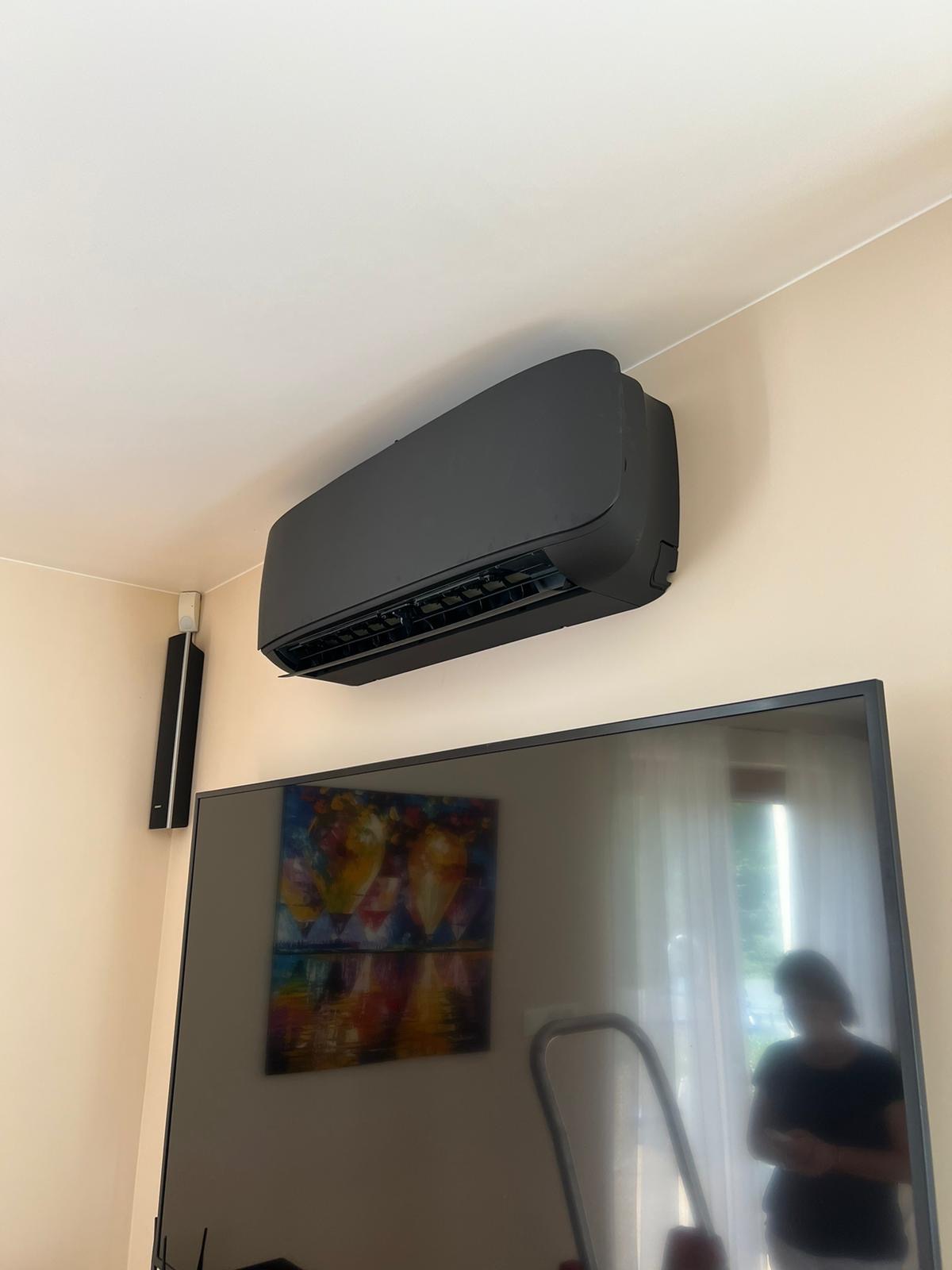 Czarny klimatyzator ścienny w nowoczesnym wnętrzu, zamontowany nad telewizorem. Widoczny fragment głośnika i obraz na ścianie. Minimalistyczny design.