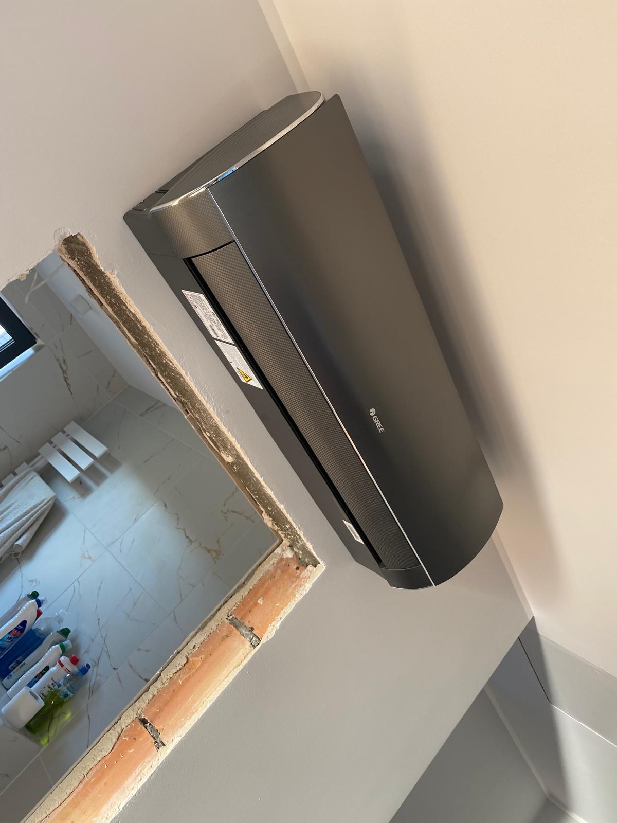 Grafitowy klimatyzator Gree zamontowany na ścianie, widoczny fragment otworu w ścianie. Ujęcie z dołu, podkreślające nowoczesny design urządzenia.