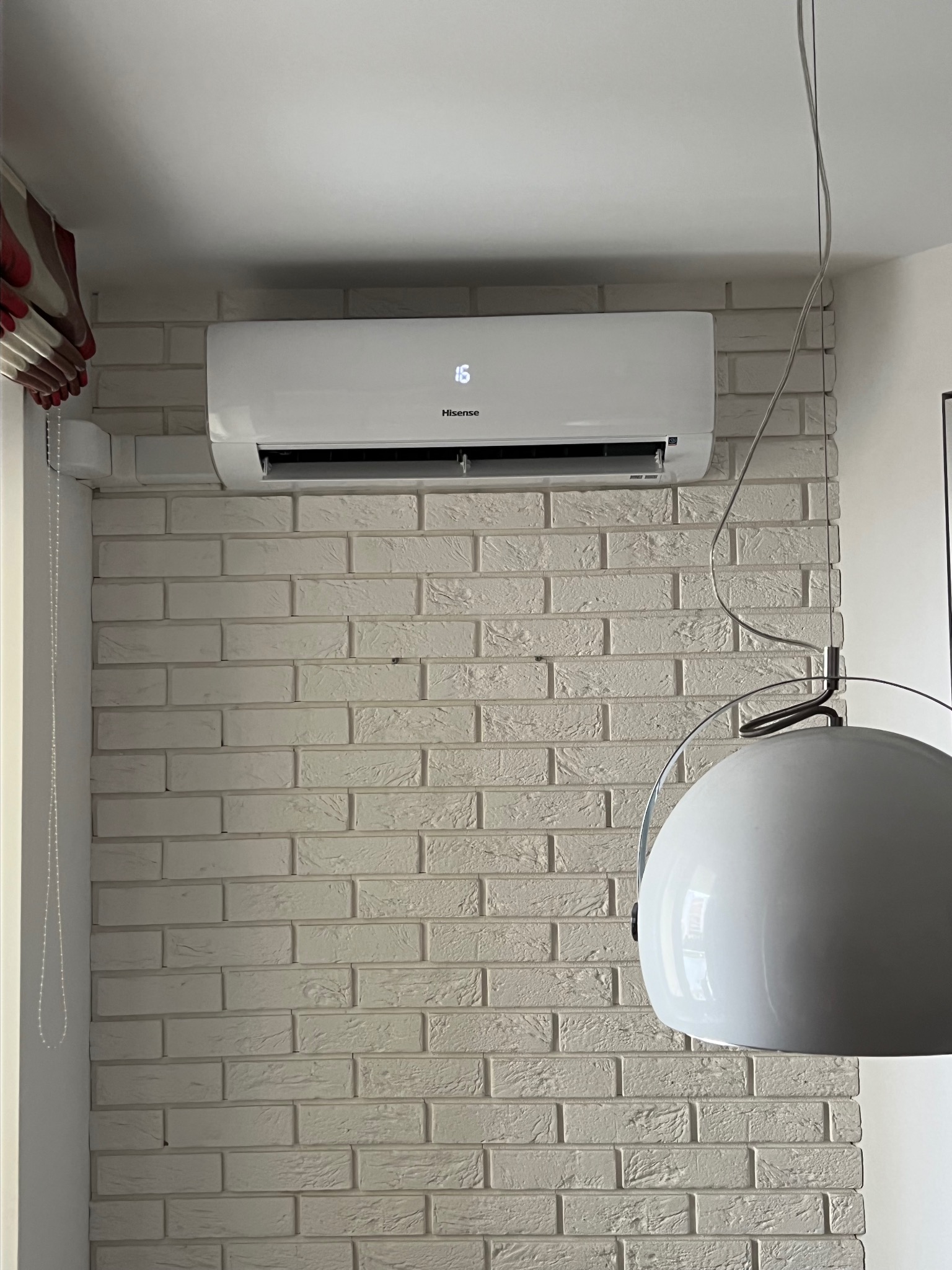 Biały klimatyzator Hisense zamontowany na ścianie z białej cegły. Widoczny fragment lampy wiszącej. Ustawiona temperatura 16 stopni.