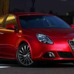 Czerwona Alfa Romeo Giulietta na tle miejskiego krajobrazu, z widoczną tablicą rejestracyjną i charakterystycznym grillem.