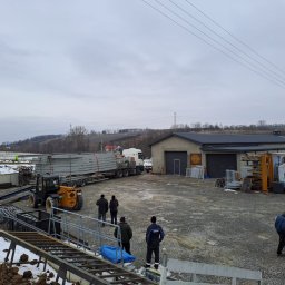 CBL Technic - Transport i rozładunek konstrukcji stalowych w Bochni. Na placu budowy widoczny wózek widłowy i pracownicy. W tle hala z bramami garażowymi i zalesione wzgórza.