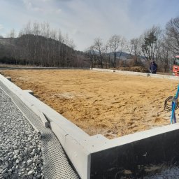 CBL Technic - Fundament pod budynek w trakcie budowy, z widoczną izolacją i podsypką piaskową. W tle drzewa i wzgórza, a w oddali sylwetka osoby.