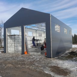 CBL Technic - Wznoszenie hali: pracownik na podnośniku montuje elementy konstrukcji. Widoczne stalowe profile, szare panele ścienne i rusztowania. Zimowy krajobraz w tle.