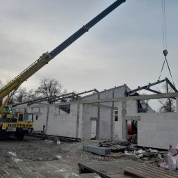 CBL Technic - Budowa hali: Żuraw podnosi stalową konstrukcję dachu na budynek z białych bloczków. Widoczny operator na podnośniku. Szaro-bura sceneria placu budowy.