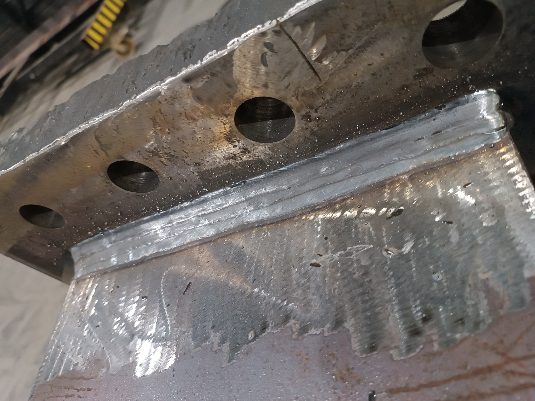 Spoina spawalnicza łącząca dwie metalowe płyty, widoczne otwory w górnej płycie. Szlifowana powierzchnia metalu. Detal konstrukcji stalowej po spawaniu.