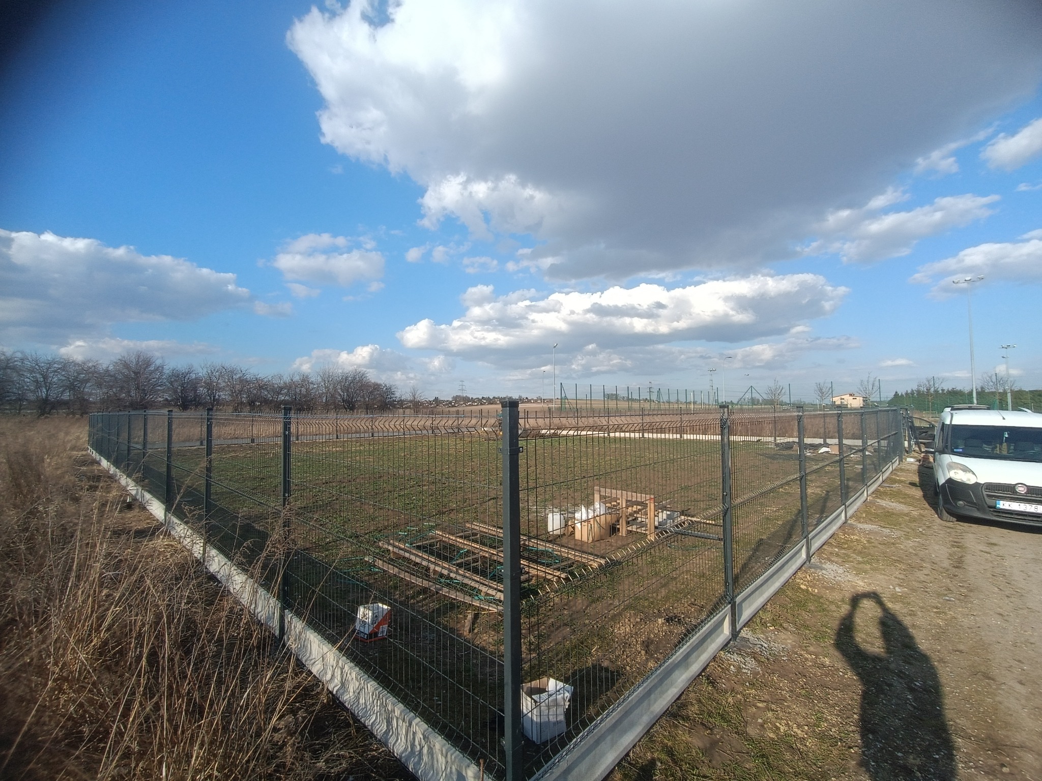 Nowoczesne ogrodzenie panelowe z podmurówką betonową otacza teren. Widoczne elementy konstrukcyjne wewnątrz ogrodzonego obszaru i zaparkowany samochód dostawczy.