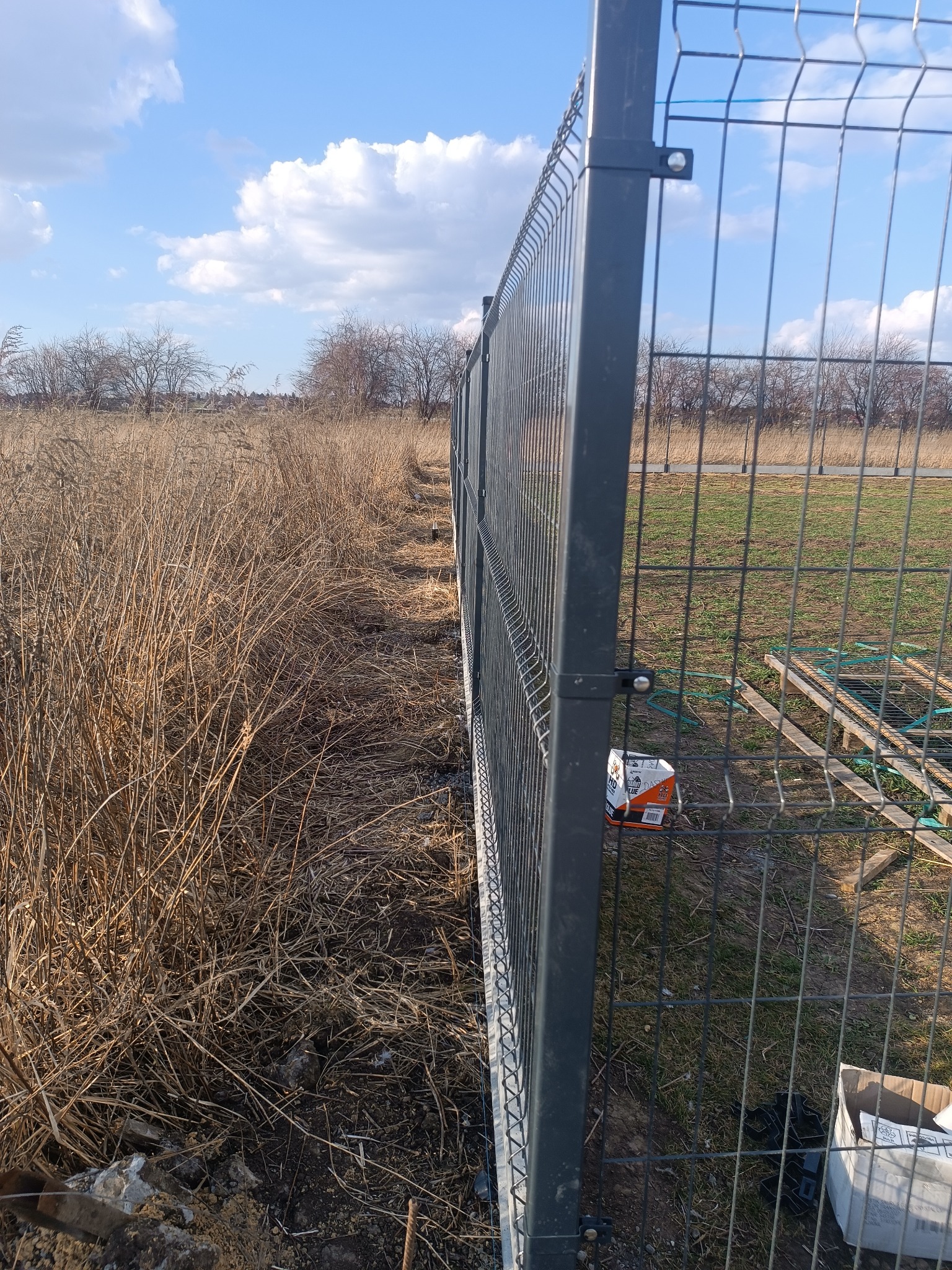 Szary panel ogrodzeniowy z siatki, widok z boku, z elementami montażowymi. W tle trawa, drzewa i błękitne niebo z chmurami. Na ziemi kartonowe pudełka.