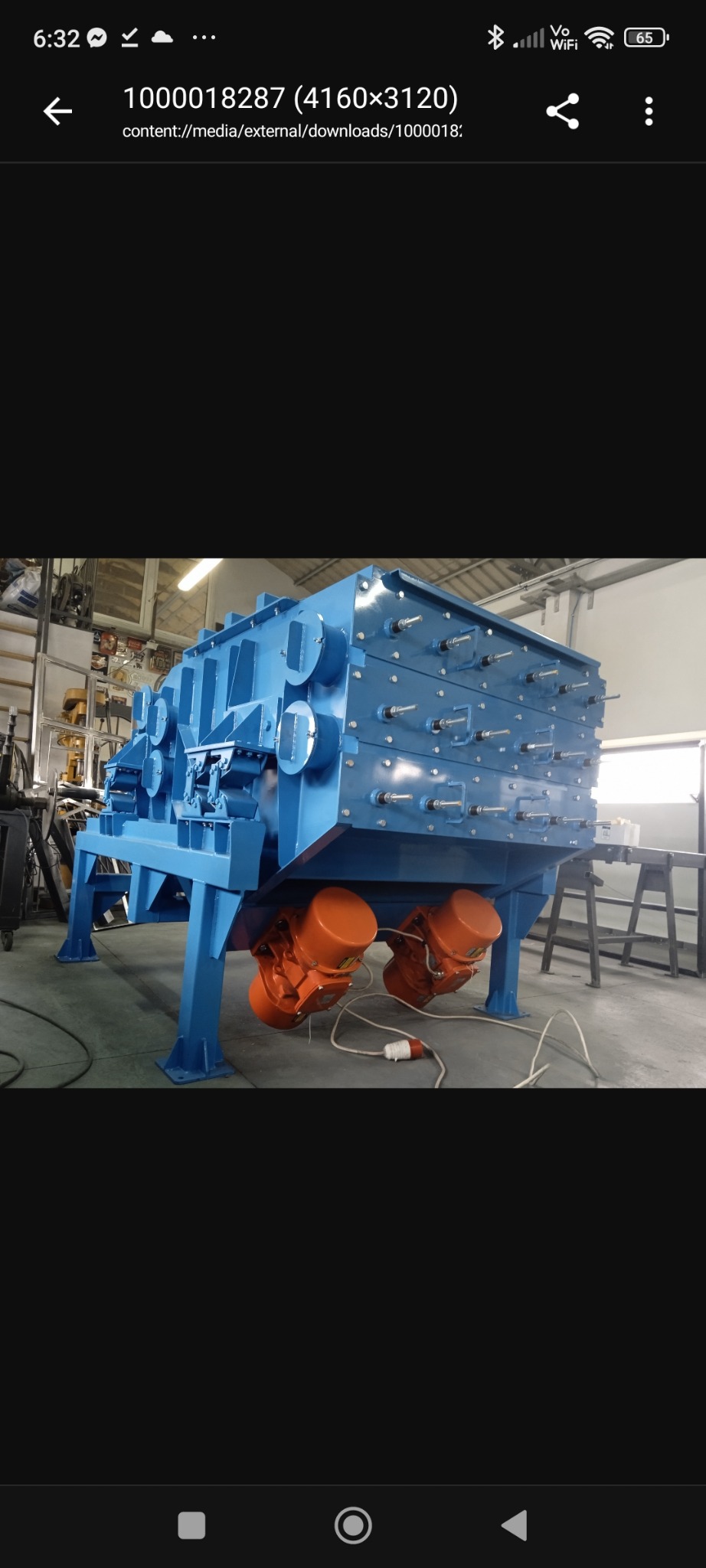 Niebieska przesiewacz wibracyjny na metalowej ramie z dwoma pomarańczowymi silnikami wibracyjnymi. Widoczne śruby i elementy konstrukcyjne. Hala produkcyjna w tle.