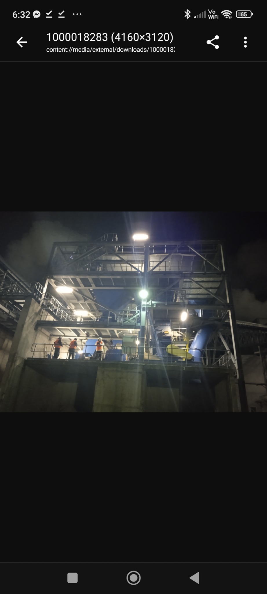 Nocne ujęcie konstrukcji stalowej hali przemysłowej z pracownikami w pomarańczowych kamizelkach, oświetlonej jasnym światłem, z widocznymi elementami instalacji.