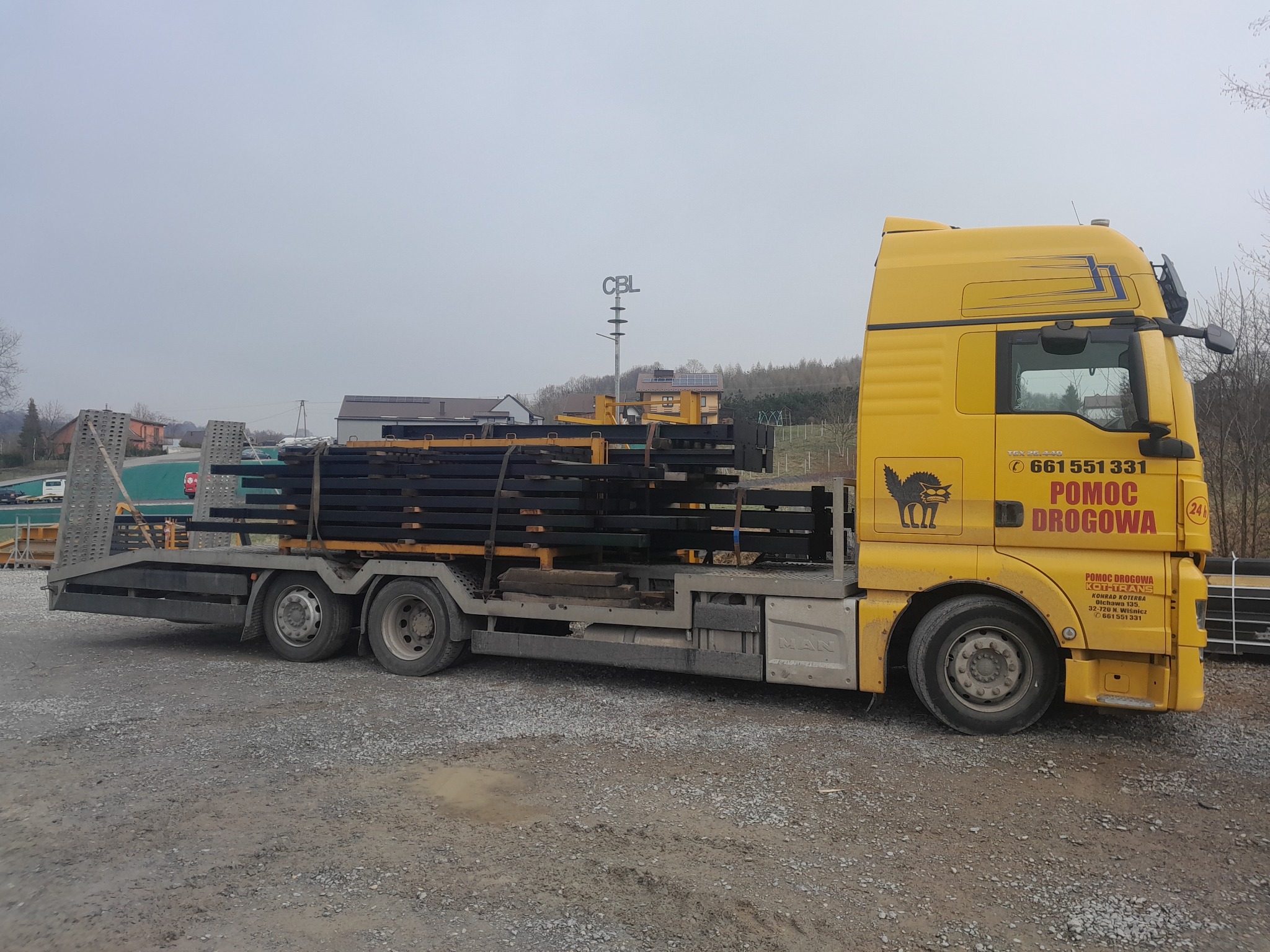 Żółta laweta MAN z czarnymi, metalowymi elementami konstrukcyjnymi na platformie, na szarym żwirowym podłożu. W tle zabudowania i słup z napisem CBL.