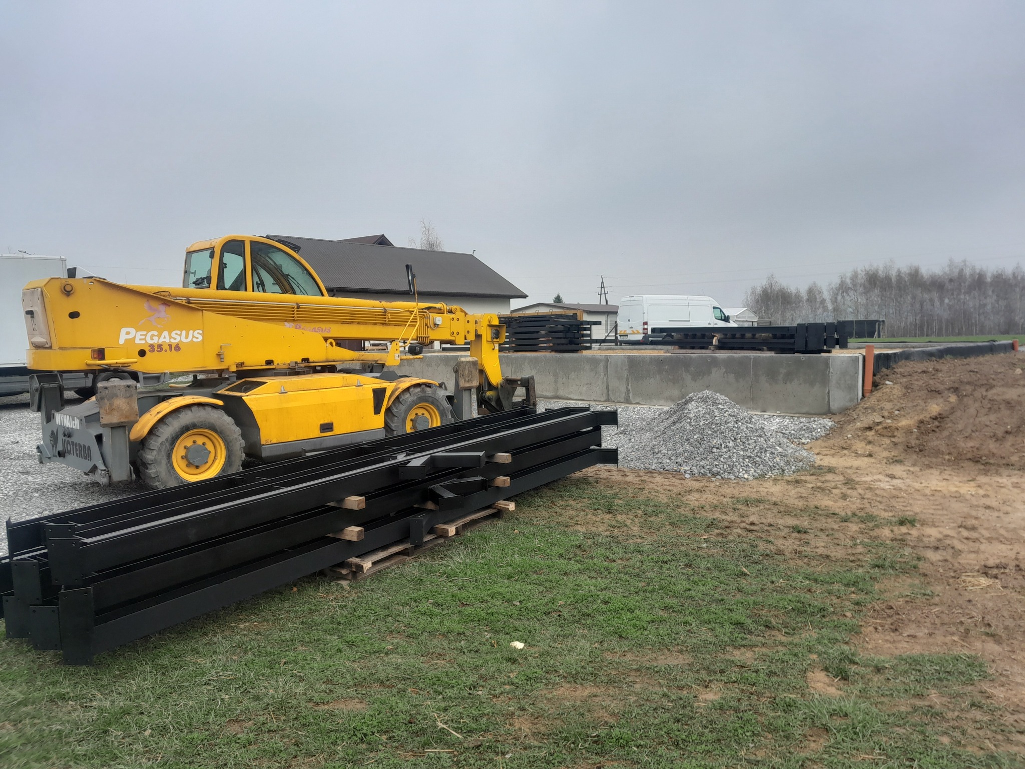 Żółty podnośnik teleskopowy Pegasus i czarne, metalowe profile na palecie, przygotowane do budowy hali na betonowym fundamencie. W tle budynek i biały bus.