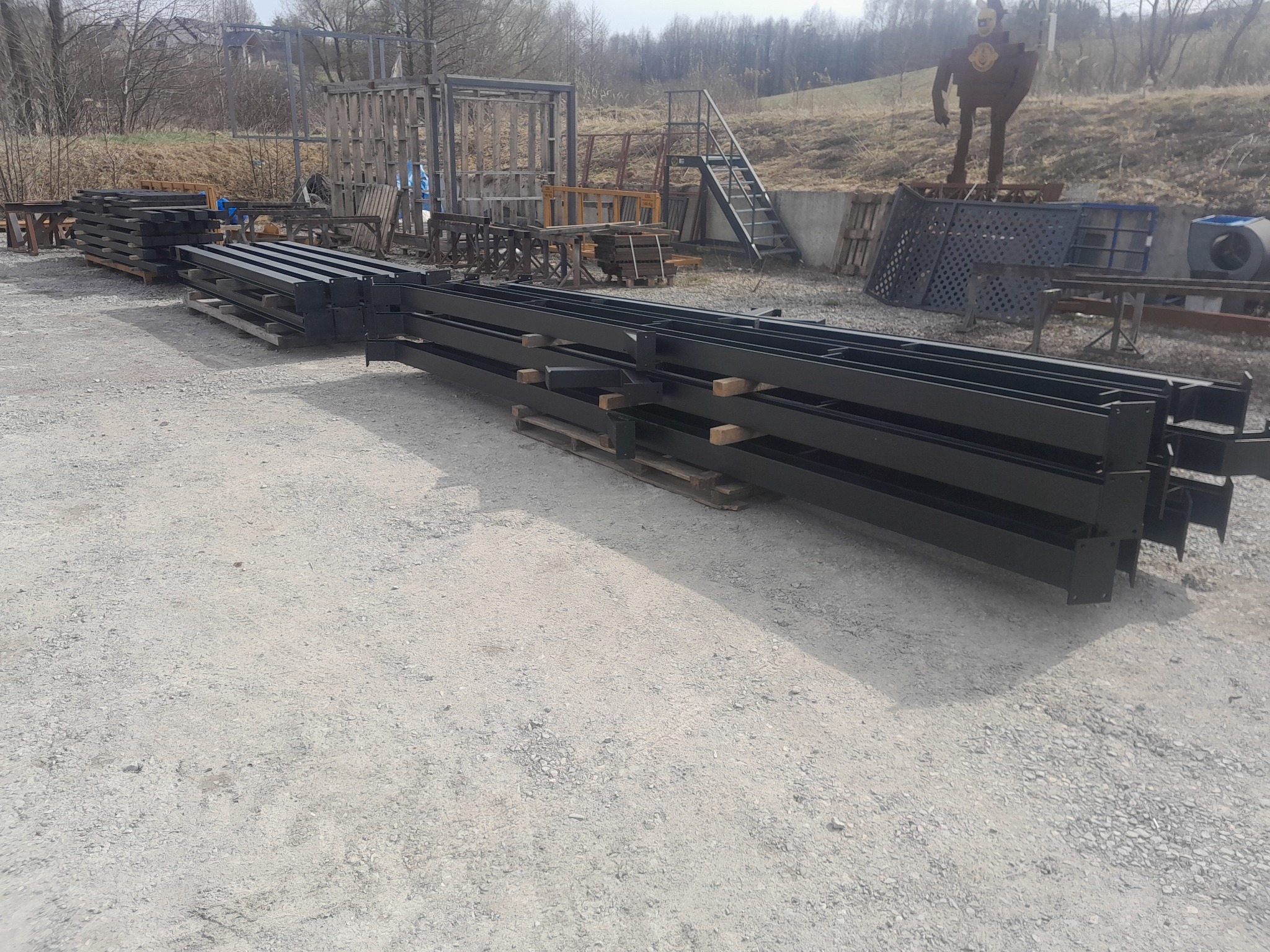 Czarne, metalowe profile na paletach, przygotowane do budowy konstrukcji stalowej. W tle widoczne elementy konstrukcyjne i metalowa rzeźba.