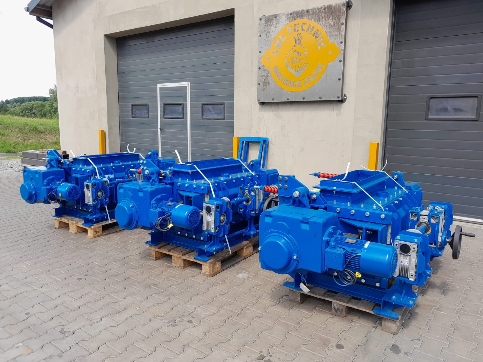 Trzy niebieskie, identyczne maszyny przemysłowe na paletach, ustawione przed szarą halą z bramami. Widoczny znak firmowy na budynku. Maszyny zabezpieczone białym sznurkiem.