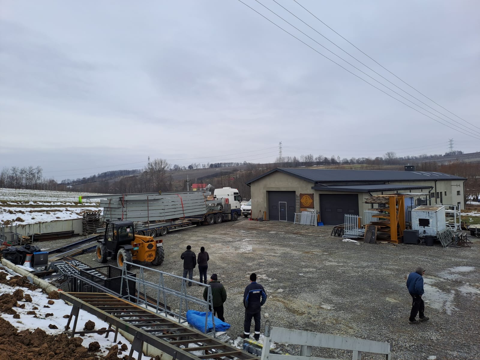 Transport i rozładunek konstrukcji stalowych w Bochni. Na placu budowy widoczny wózek widłowy i pracownicy. W tle hala z bramami garażowymi i zalesione wzgórza.