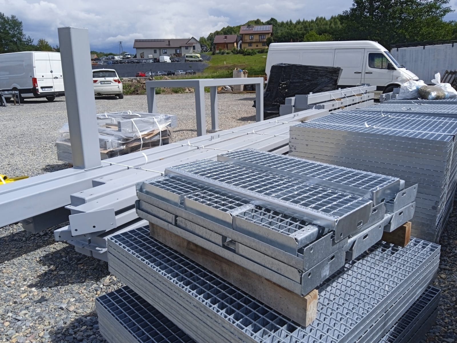 Stalowe elementy konstrukcyjne i kraty pomostowe w szarym kolorze, przygotowane do montażu na placu budowy. W tle budynki i zaparkowane samochody.