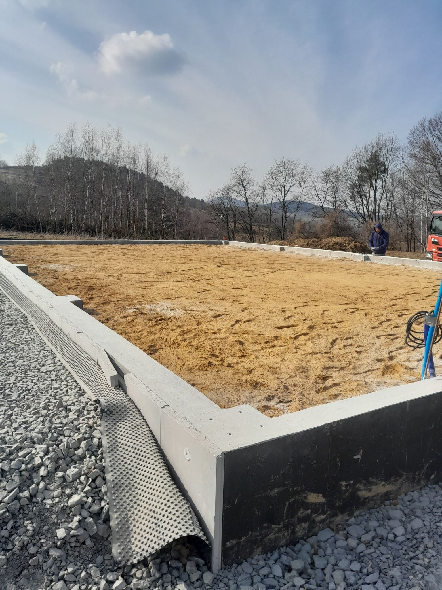 Fundament pod budynek w trakcie budowy, z widoczną izolacją i podsypką piaskową. W tle drzewa i wzgórza, a w oddali sylwetka osoby.