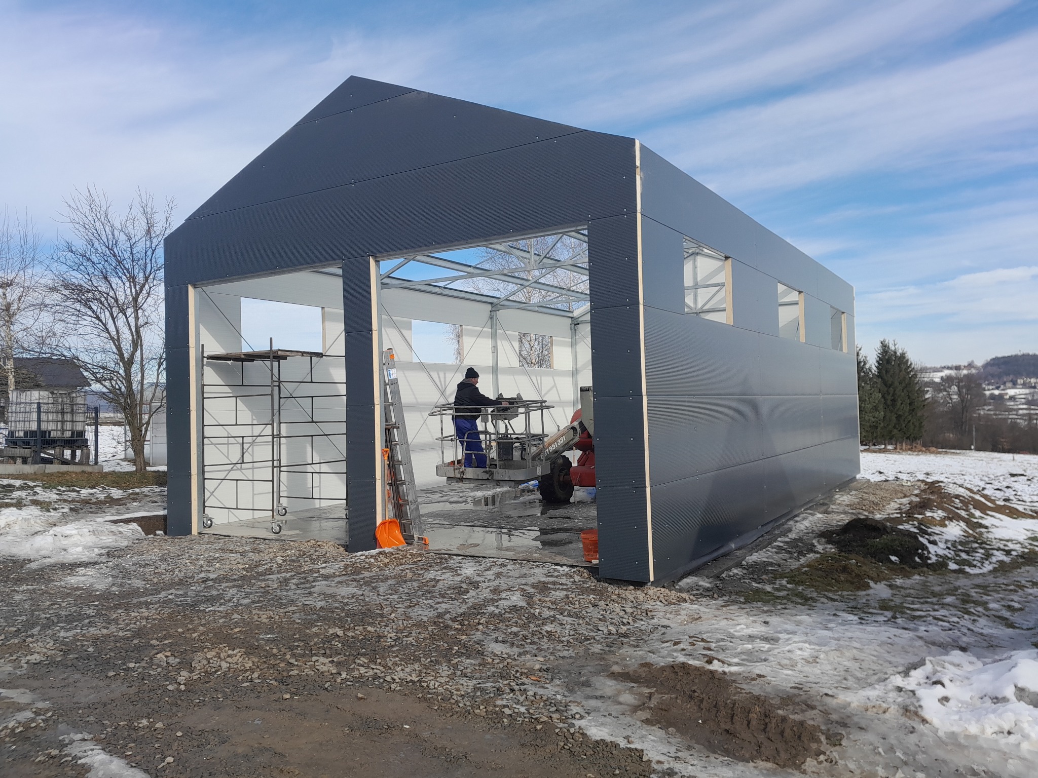 Wznoszenie hali: pracownik na podnośniku montuje elementy konstrukcji. Widoczne stalowe profile, szare panele ścienne i rusztowania. Zimowy krajobraz w tle.