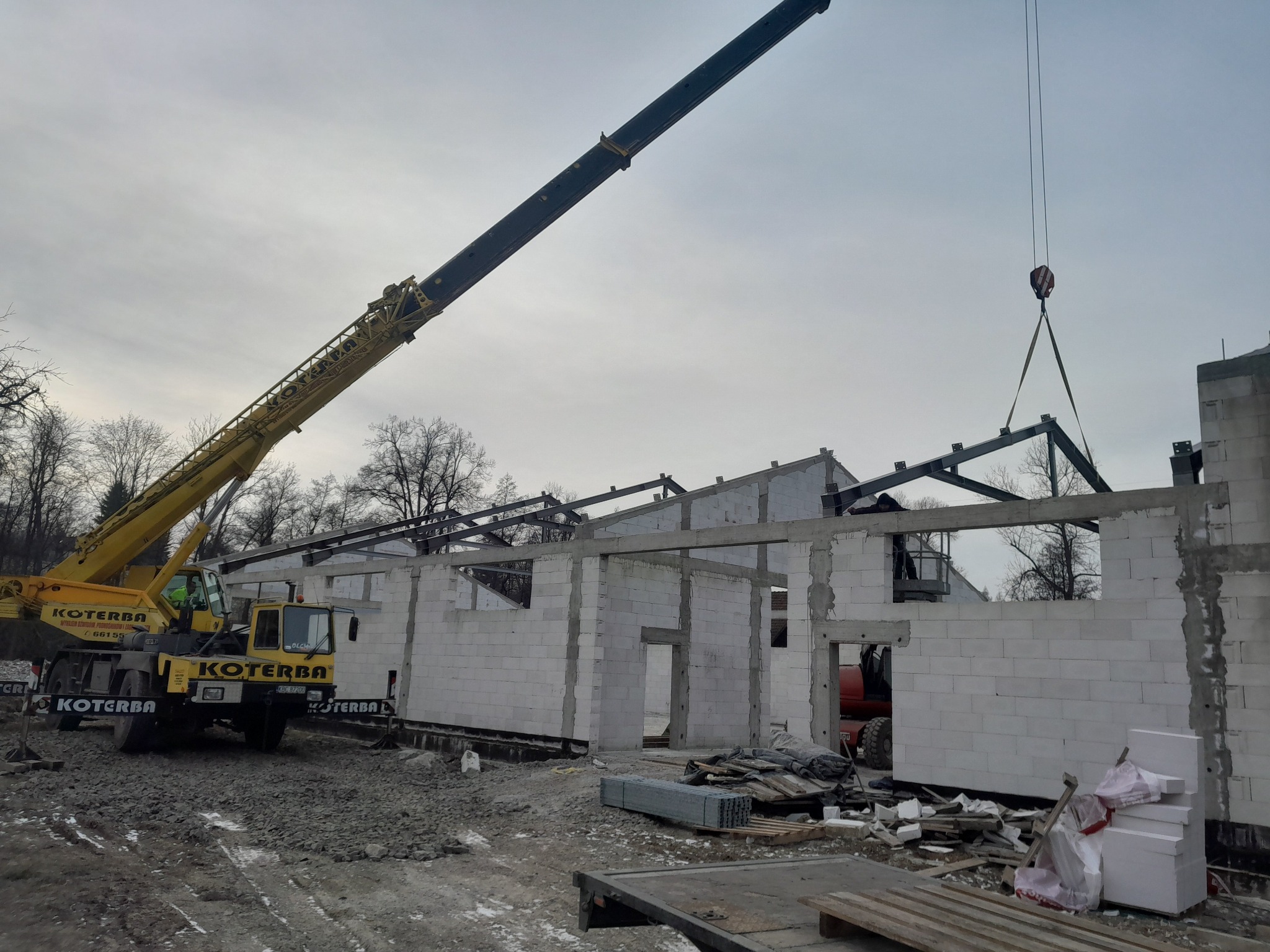 Budowa hali: Żuraw podnosi stalową konstrukcję dachu na budynek z białych bloczków. Widoczny operator na podnośniku. Szaro-bura sceneria placu budowy.