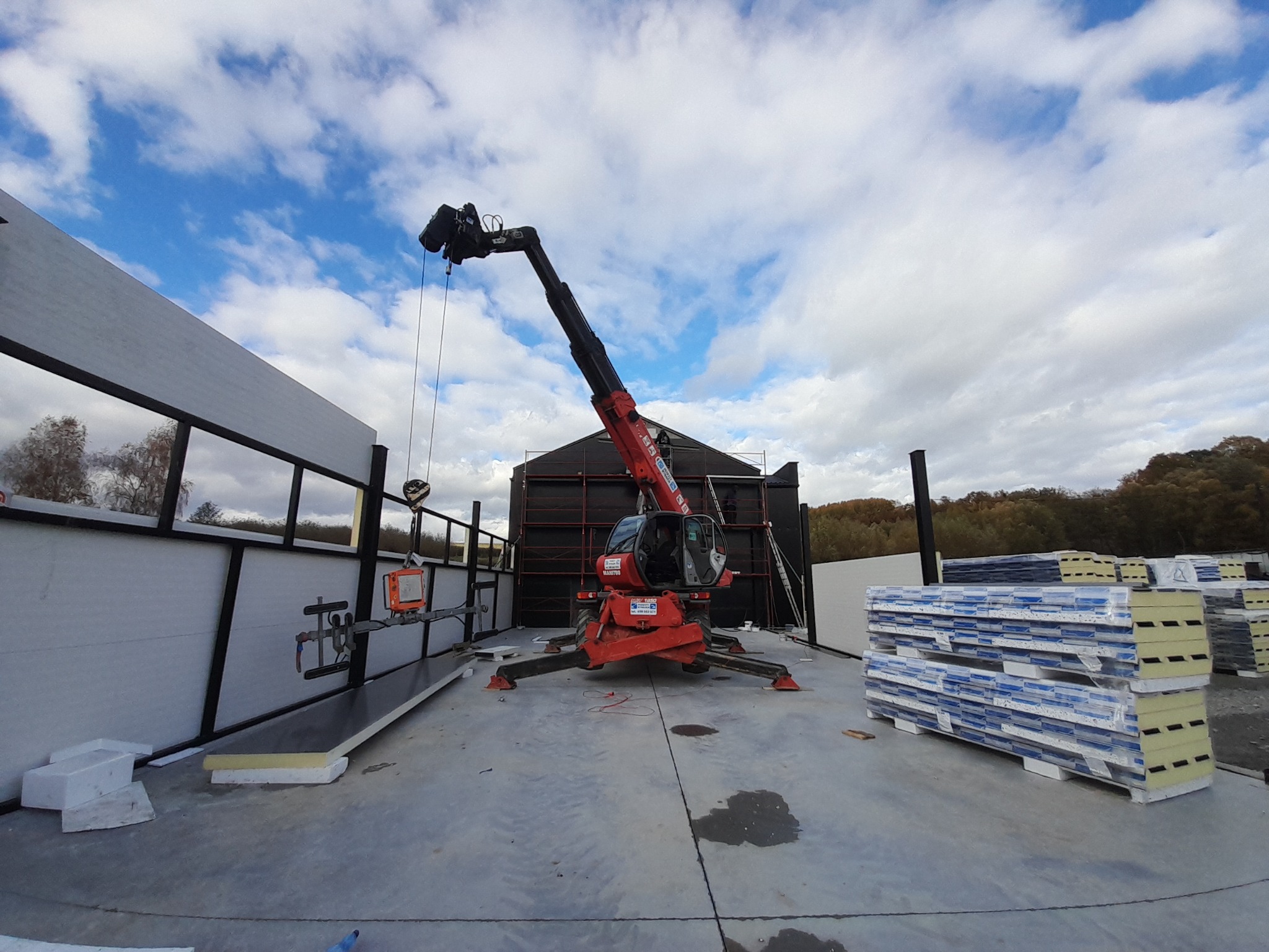 Montaż paneli warstwowych hali przemysłowej za pomocą podnośnika teleskopowego. Widoczne elementy konstrukcji i przygotowane panele. Pochmurne niebo w tle.