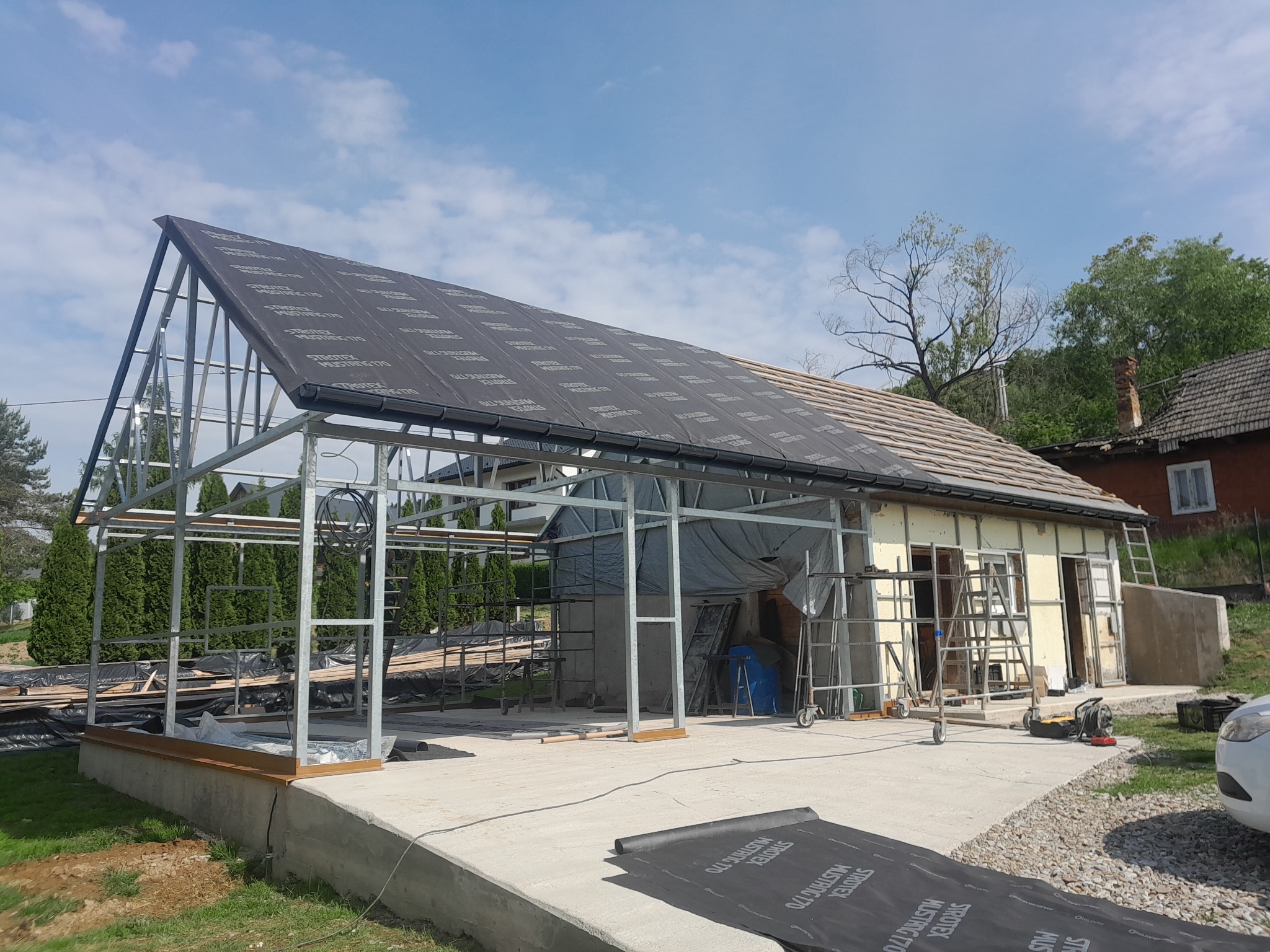 Konstrukcja stalowa hali w trakcie budowy, widoczne elementy dachu pokryte membraną, rusztowania i narzędzia budowlane. Częściowo ocieplona ściana budynku.