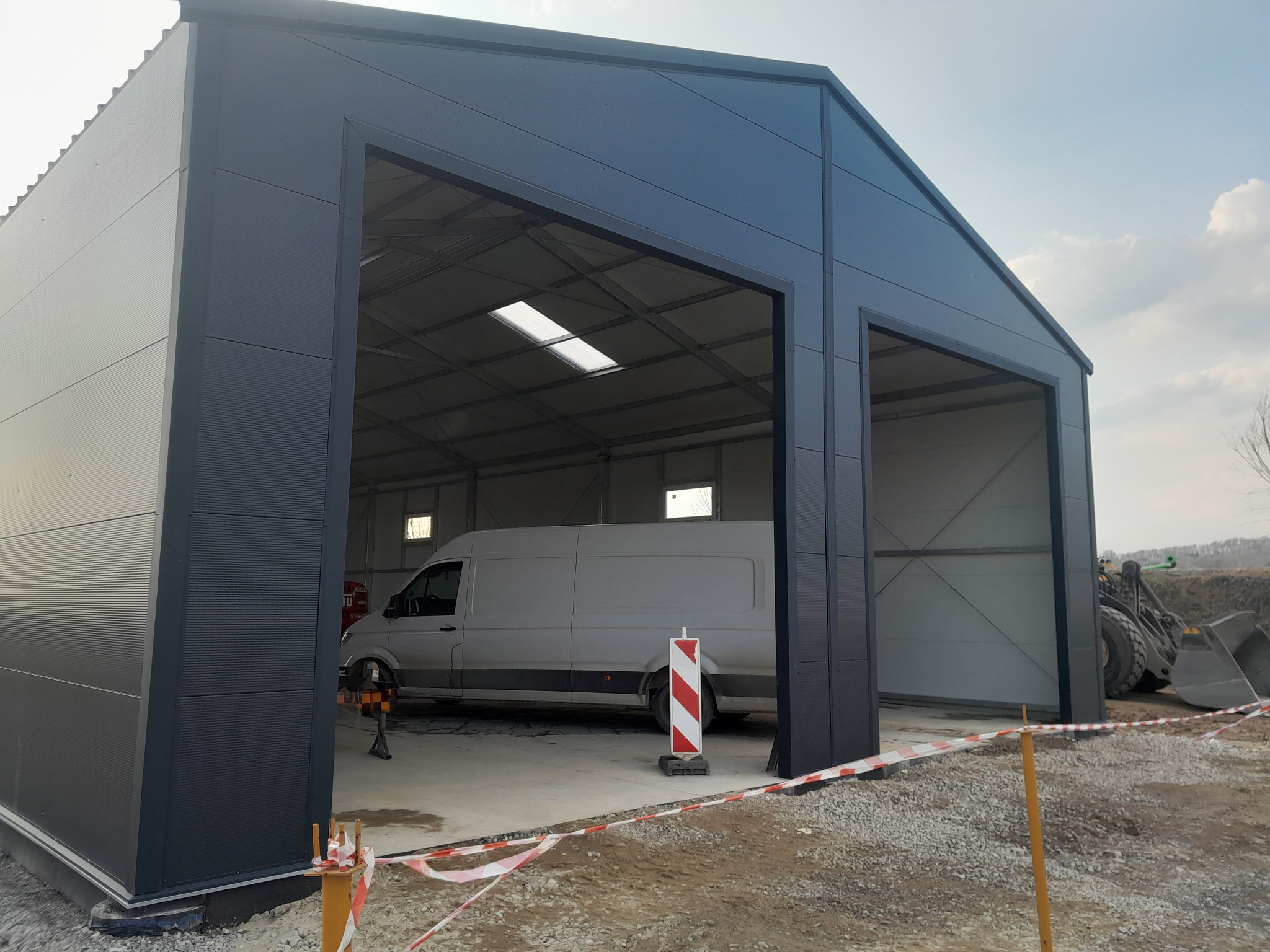 Nowoczesna hala stalowa z dwoma wjazdami, wewnątrz biały bus. Zewnętrzne wykończenie w kolorze grafitowym, widoczne elementy konstrukcyjne i świetliki dachowe.