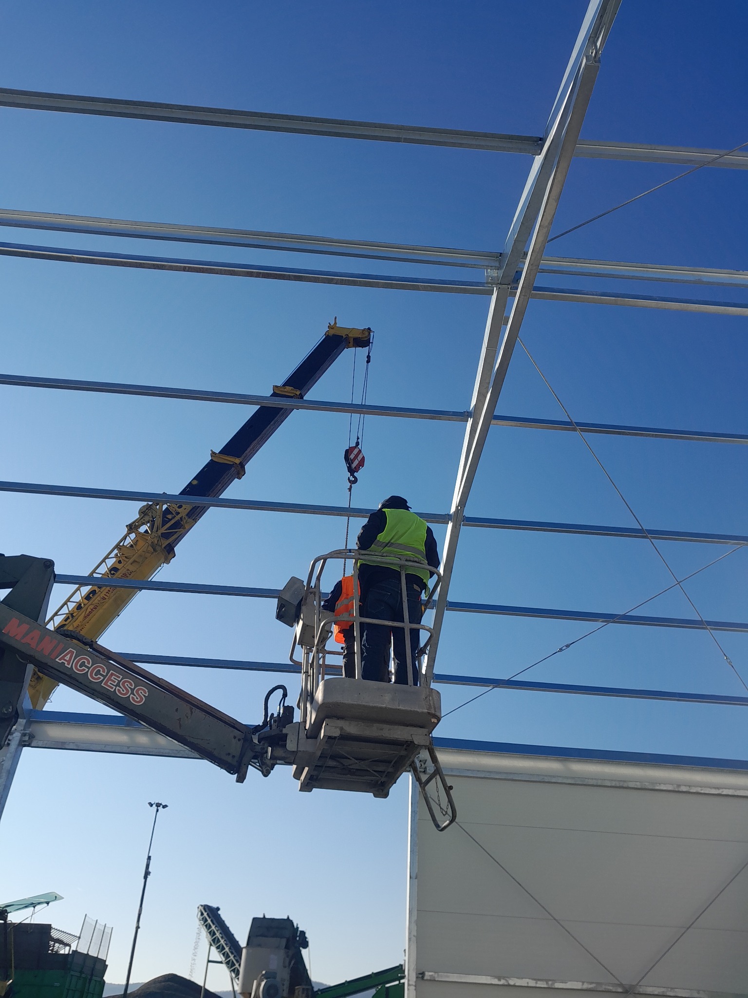 Pracownicy na podnośniku montują stalową konstrukcję hali na tle błękitnego nieba. Widoczny dźwig i elementy konstrukcyjne. Etap budowy z użyciem specjalistycznego sprzętu.