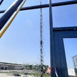 ProMaClean Maciej Wąsek - Mycie okna z zewnątrz za pomocą szczotki na teleskopowym kiju. Woda spływa po szybie, a w tle widoczne budynki i niebo.