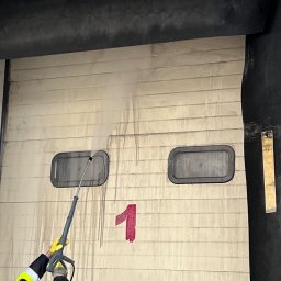 ProMaClean Maciej Wąsek - Czyszczenie bramy garażowej z numerem '1' myjką ciśnieniową. Widoczne ślady brudu spływające po powierzchni. Osoba w żółtych rękawicach obsługuje urządzenie.