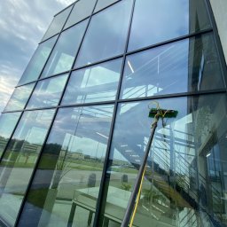 ProMaClean Maciej Wąsek - Czyszczenie szklanej fasady biurowca z użyciem lancy teleskopowej. Widoczne smugi wody spływające po szybie. W tle odbicie nieba i wnętrza budynku.