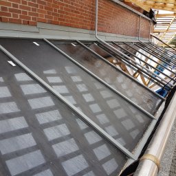 ProMaClean Maciej Wąsek - Ukośny widok na umyte, przeszklone panele dachowe z aluminiową ramą, mokre od wody, z widoczną ceglaną ścianą i elementami konstrukcji dachu.