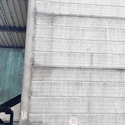 ProMaClean Maciej Wąsek - Szara, pozioma elewacja z blachy falistej, z widocznymi zabrudzeniami i śladami po myciu. Fragment dachu i betonowej podstawy w kadrze.