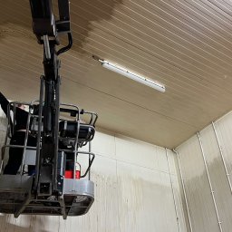 ProMaClean Maciej Wąsek - Pracownik w podnośniku koszowym czyści sufit i ściany hali przemysłowej. Widoczne ślady czyszczenia, oświetlenie jarzeniowe i elementy konstrukcyjne budynku.