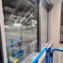 ProMaClean Maciej Wąsek - Okno hali magazynowej widziane z podnośnika koszowego. Widoczne odbicie w szybie i fragment konstrukcji podnośnika.