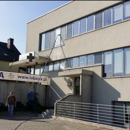 ProMaClean Maciej Wąsek - Czyszczenie okien na wysokości w budynku apteki. Widoczny pracownik z lancą teleskopową i budynek z licznymi oknami. Ujęcie z dołu, w tle budynek mieszkalny.