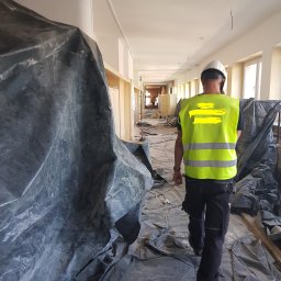 ProMaClean Maciej Wąsek - Pracownik w kasku i kamizelce odblaskowej idzie korytarzem zabezpieczonym folią malarską po sprzątaniu po remoncie. W tle widoczne okna i resztki gruzu.