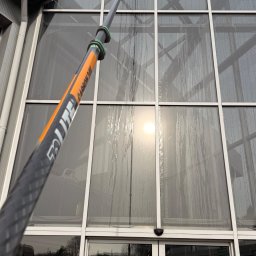 ProMaClean Maciej Wąsek - Czyszczenie przeszklonej fasady budynku za pomocą teleskopowego kija z myjką. Widoczne smugi wody na szybach i odbicie słońca. Ujęcie z dołu, ukazujące wysokość budynku.