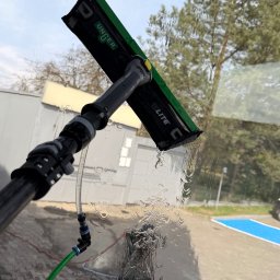 ProMaClean Maciej Wąsek - Profesjonalne mycie okna z użyciem szczotki teleskopowej Unger i węża doprowadzającego wodę. Widoczne krople wody na szybie i budynek w tle.