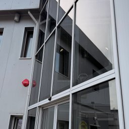 ProMaClean Maciej Wąsek - Szklana fasada budynku biurowego z widocznym odbiciem otoczenia i elementami architektonicznymi. Na ścianie znak 'Pożar'.